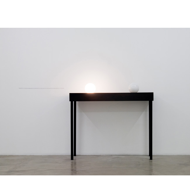 Iman Issa. <em>Material for a sculpture proposed as an alternative to a monument that has become an embarrassment to its people</em>, 2010. Two lightbulbs, plywood table, and vinyl text on the wall; 59 x 47-1/2 x 19-3/4 inches. Courtesy the artist and Rodeo Gallery, Istanbul. Iman Issa, Material for a sculpture proposed as an alternative to a monument that has become an embarrassment to its people